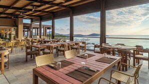 A spacious outdoor dining area with wooden tables and chairs, large windows offering a view of the sea and mountains.