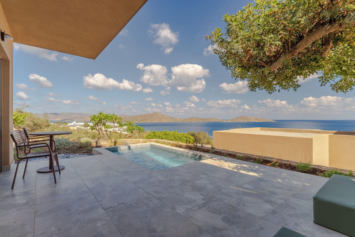 A pool area with a table and chairs, surrounded by greenery and a view of the sea and hills.