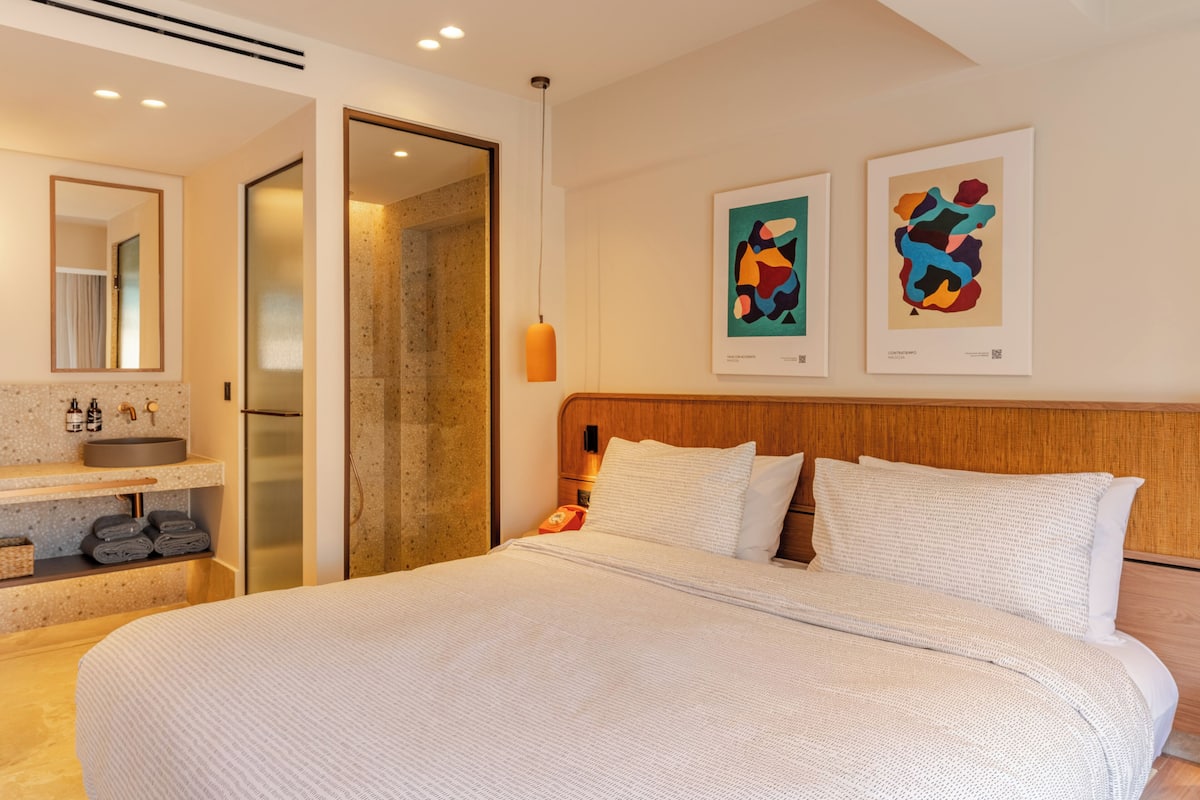 A modern hotel room with a large bed, a wooden headboard, a bathroom with a sink and mirror, and two abstract art pieces on the wall.