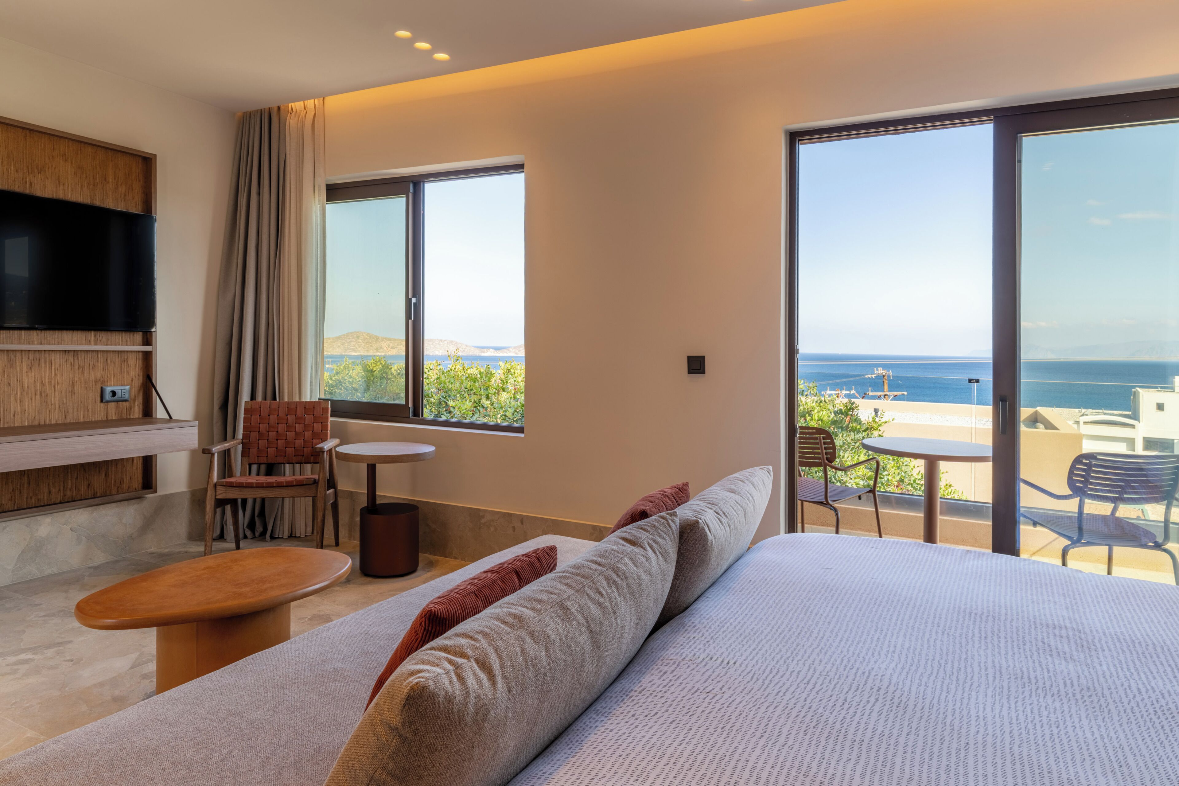 A modern hotel room with a large bed, a TV, a balcony with a view of the sea, and a seating area with chairs.