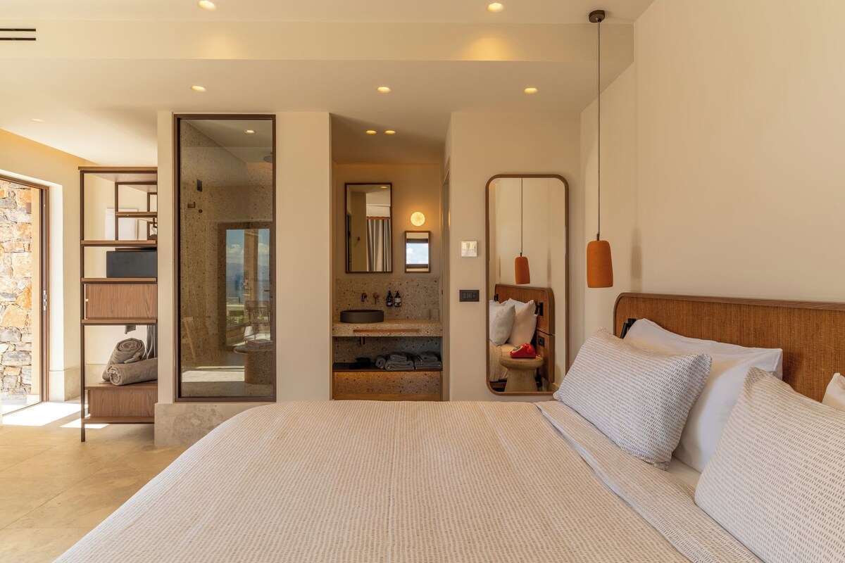 A modern hotel room with a large bed, a bathroom with a mirror, and a stone wall.