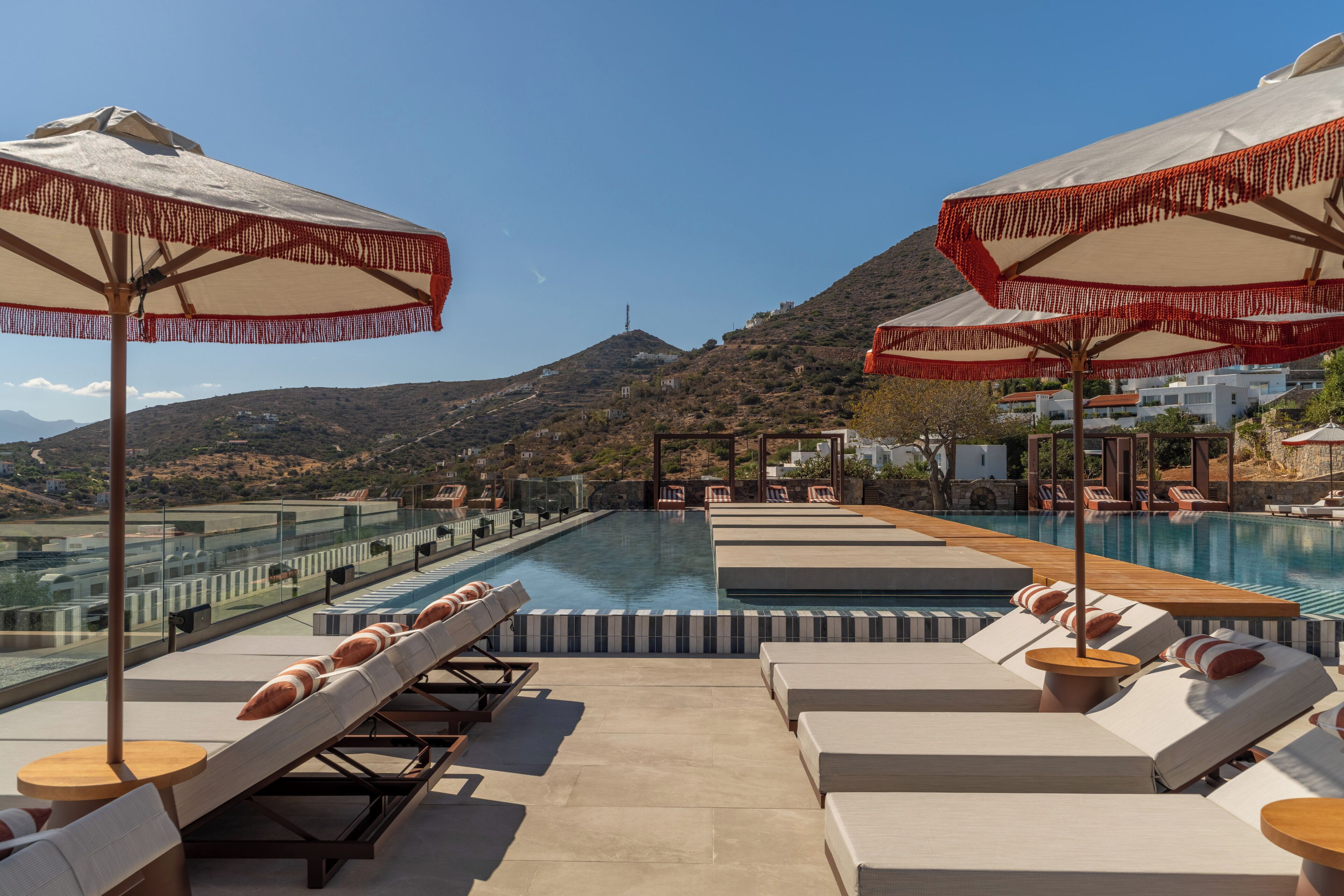 Zona junto a la piscina con tumbonas y sombrillas, vista despejada del agua y montañas al fondo.