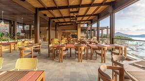 An outdoor dining area with wooden tables and chairs, large windows offering a view of the sea and boats.