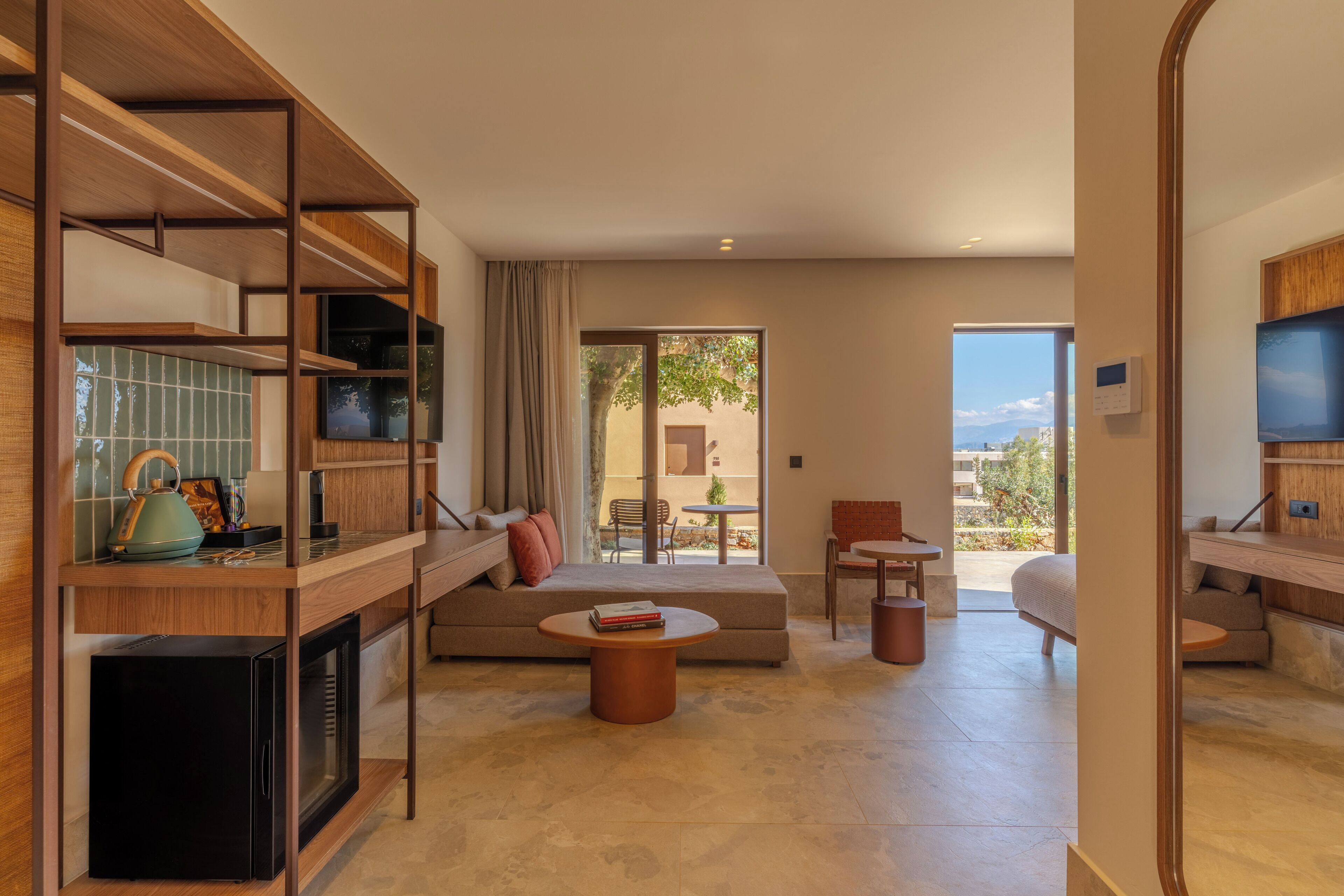 A modern hotel room with a kitchen area, a seating area with a sofa and coffee table, and a balcony with a view.