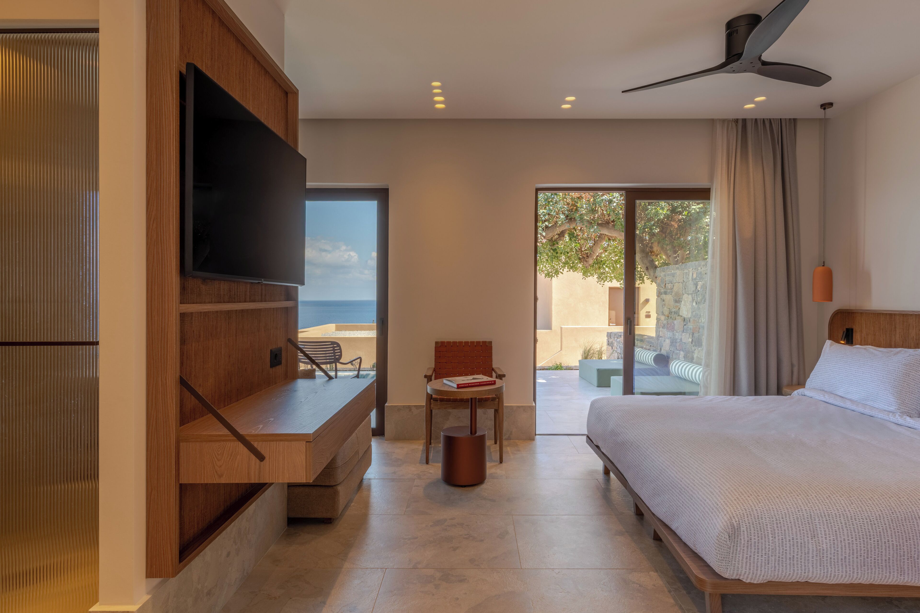 A modern hotel room with a large bed, a flat-screen TV mounted on a wooden panel, a small table with a chair, and a view of the outdoors through a glass door.