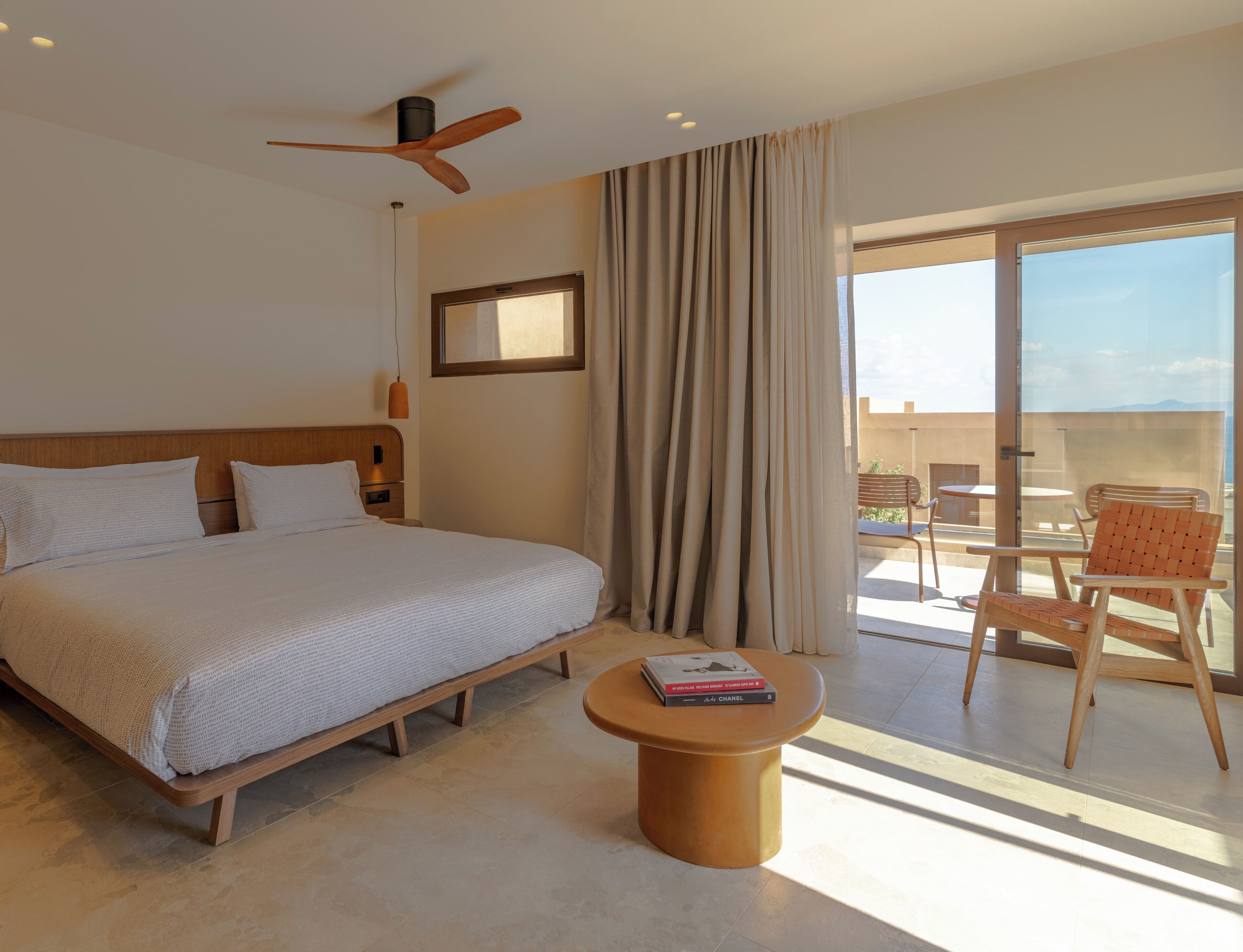 A hotel room with a bed, a round table, a chair, a ceiling fan, and a view of the outdoors.