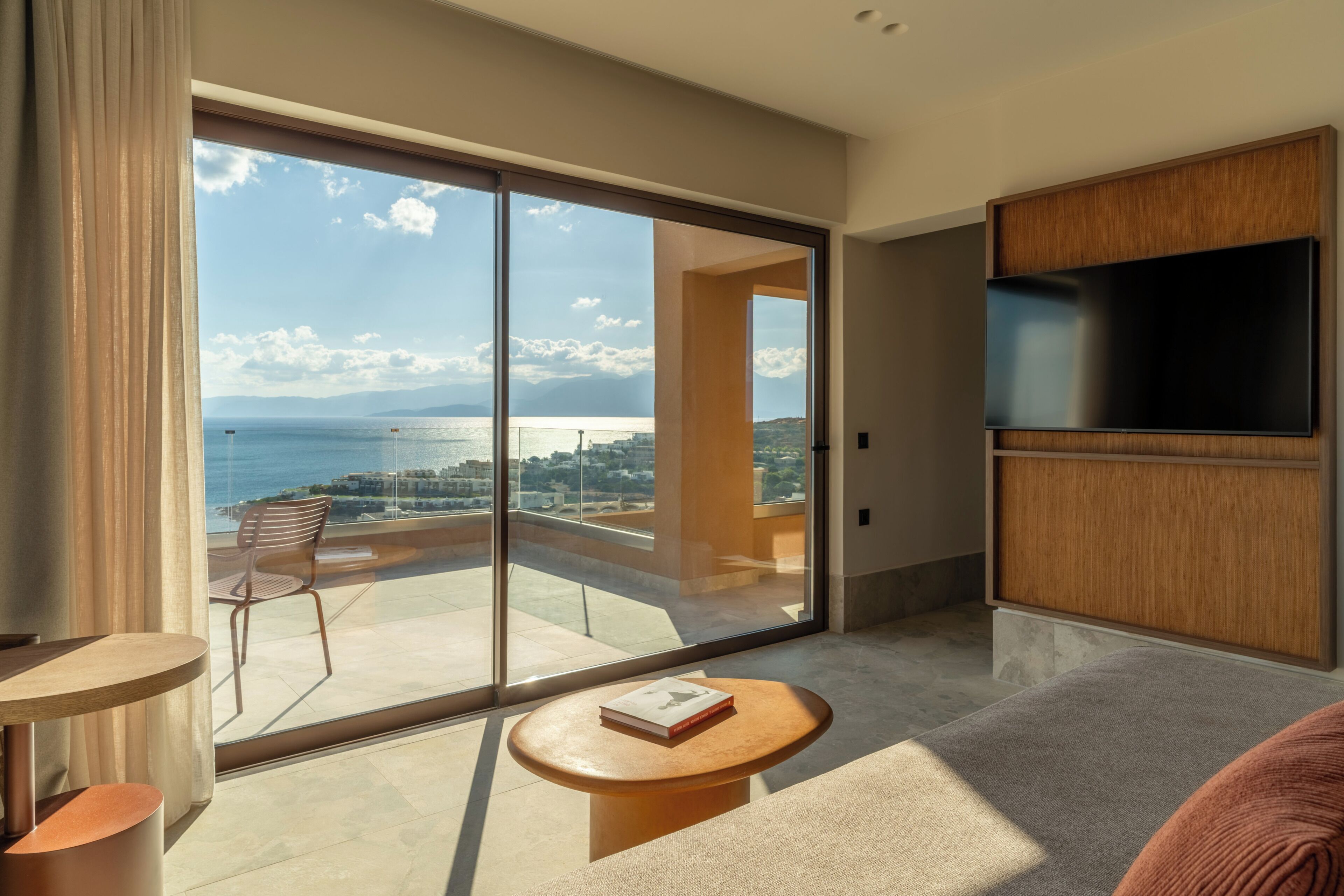 A modern hotel room with a large sliding glass door to a balcony, a flat-screen TV, and a small round table with a book.