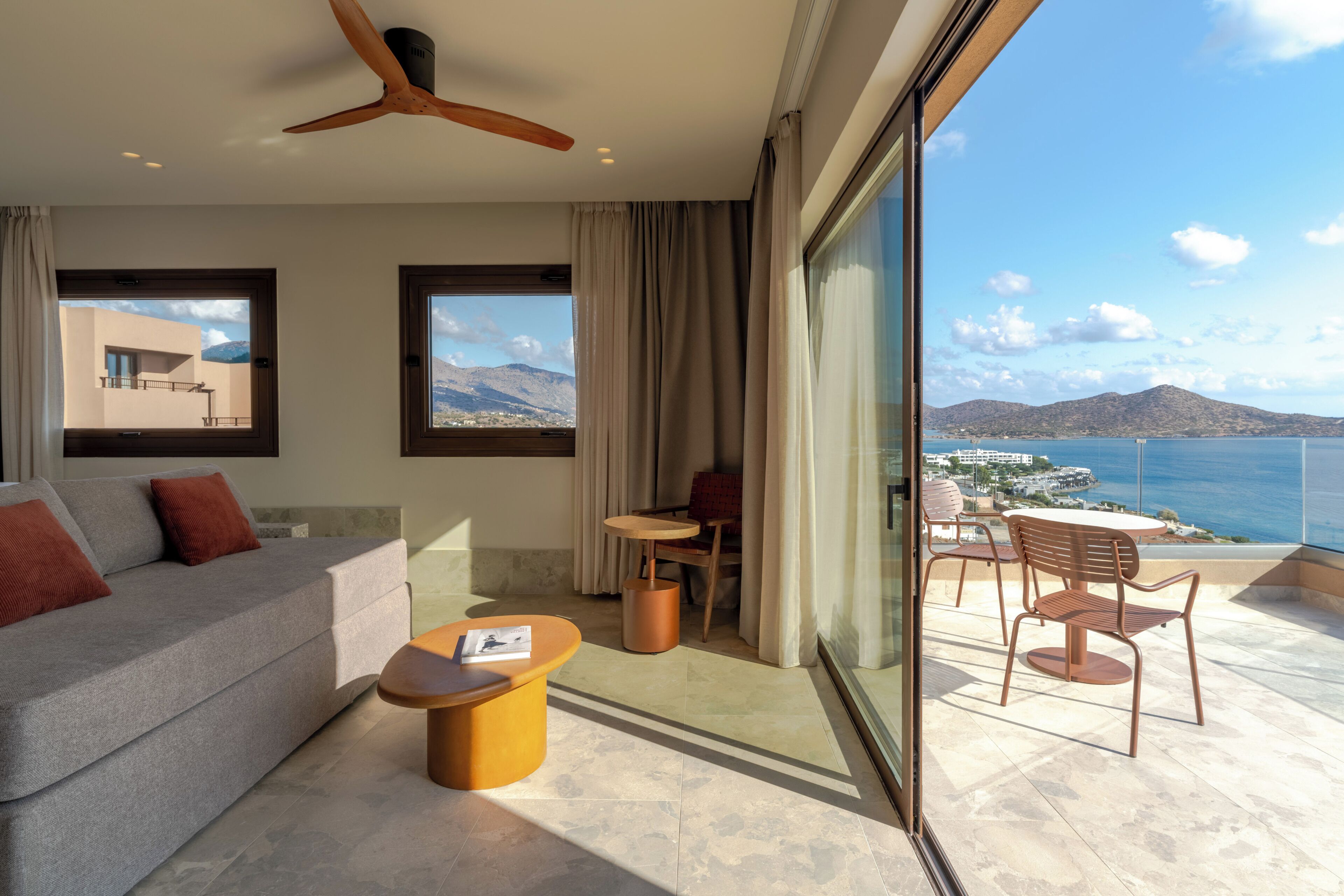 A modern hotel room with a balcony, a sofa, a coffee table, and a view of the sea and mountains.