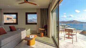A modern hotel room with a balcony, a sofa, a coffee table, and a view of the sea and mountains.
