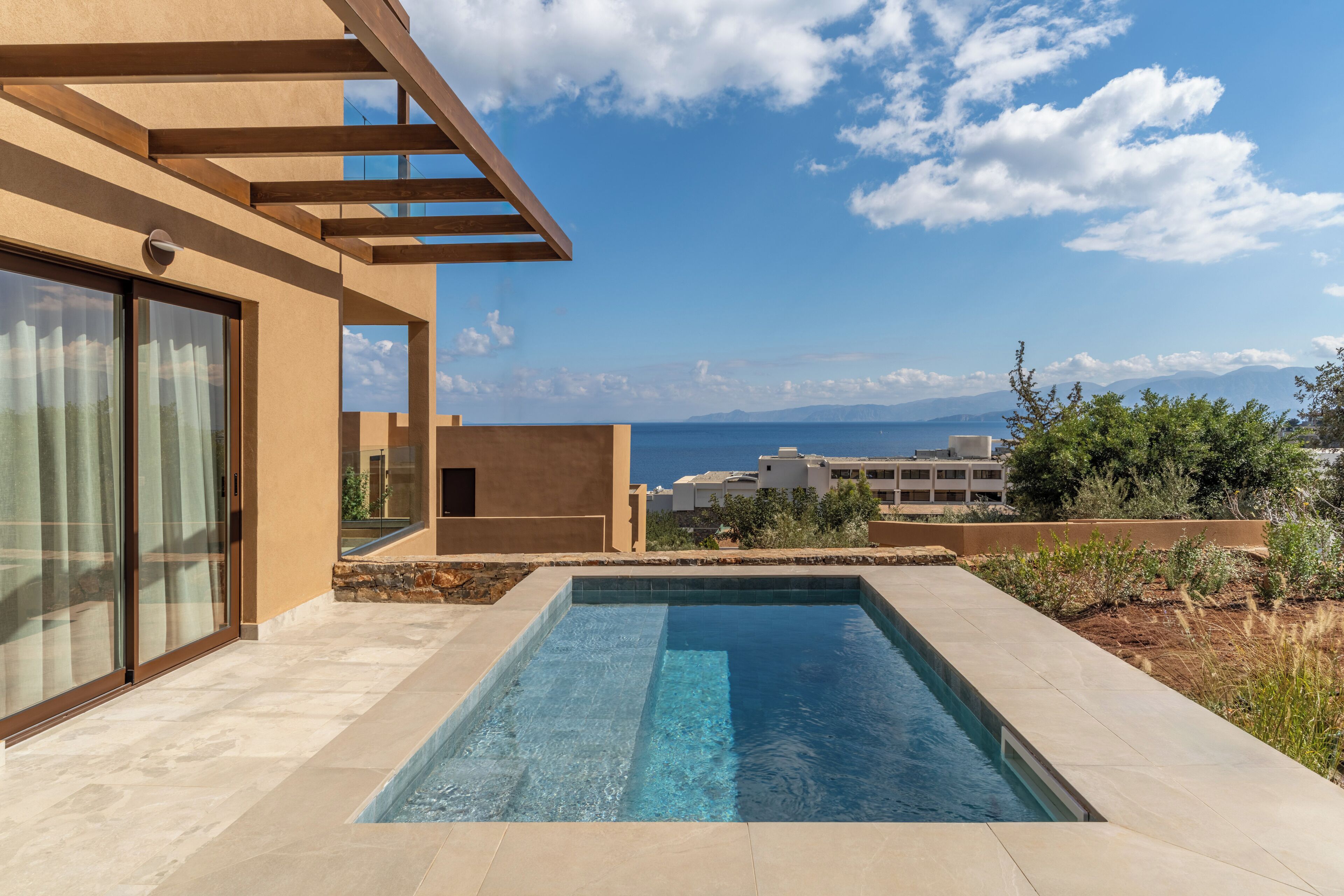 Ein moderner Poolbereich mit klarem, blauem Wasser, umgeben von einem beige gefärbten Gebäude mit großen Fenstern und einer Pergola.