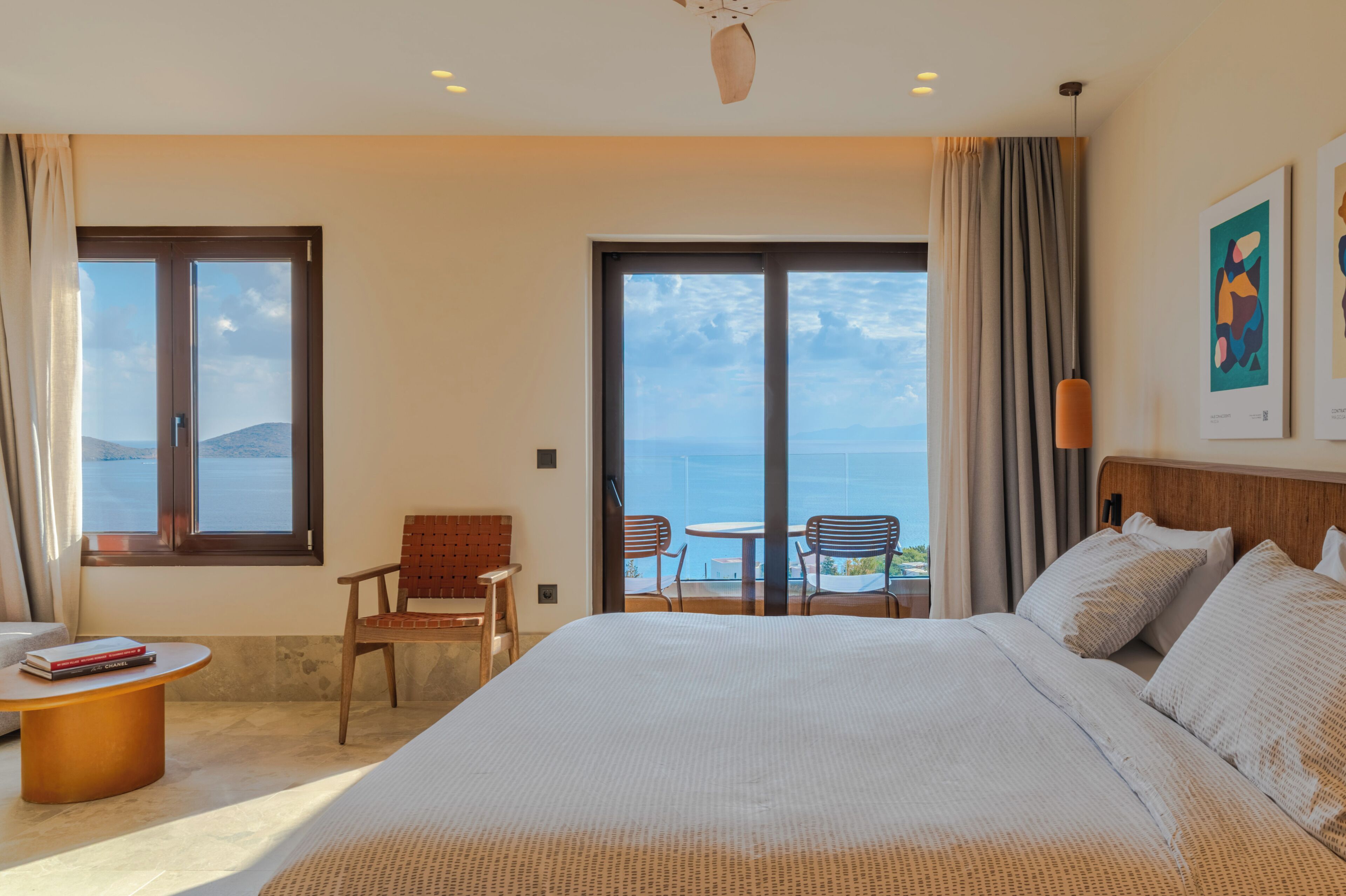 A hotel room with a large bed, a small table, two chairs, and a balcony with a view of the sea.