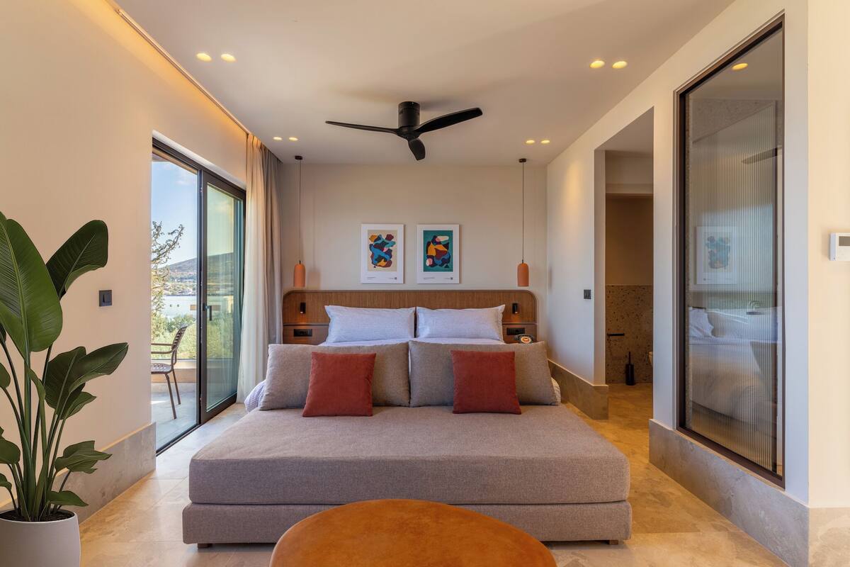 A modern bedroom with a large bed, a ceiling fan, artwork on the wall, and a balcony with a view.