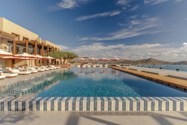 Piscina con sillones de playa y sombrillas, rodeada por un edificio con grandes ventanales y vistas a las montañas.
