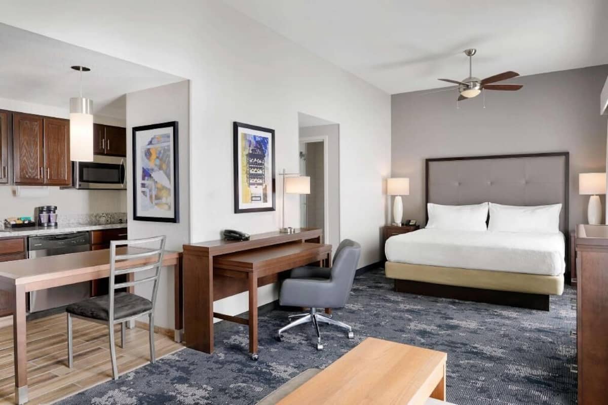 A hotel room with a bed, desk, chair, kitchenette, and ceiling fan.