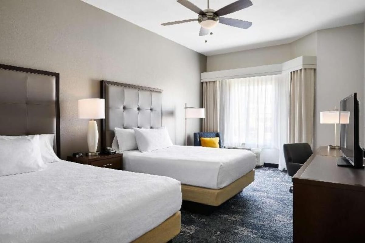 Une chambre d’hôtel avec deux lits, un bureau, une télévision, un ventilateur de plafond et une fenêtre avec des rideaux.