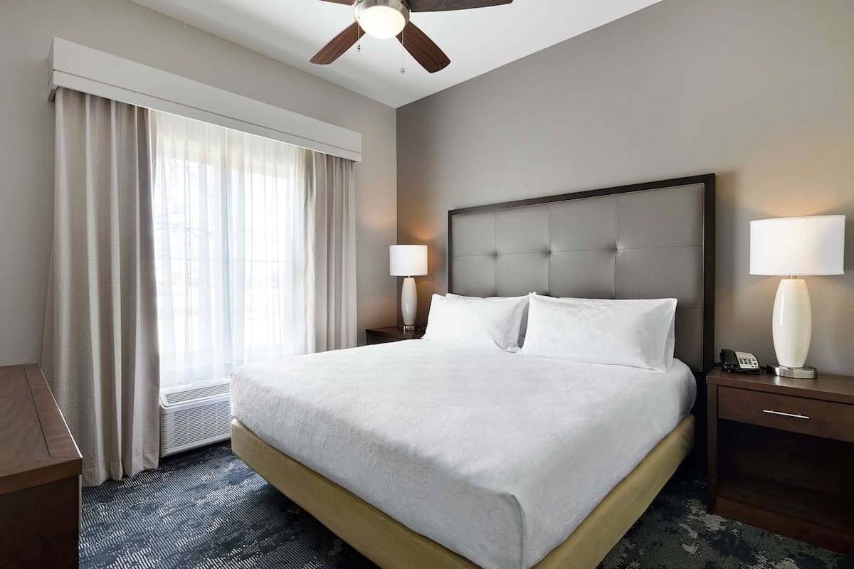 A hotel room with a large bed, a ceiling fan, a nightstand with a lamp, and a window with curtains.