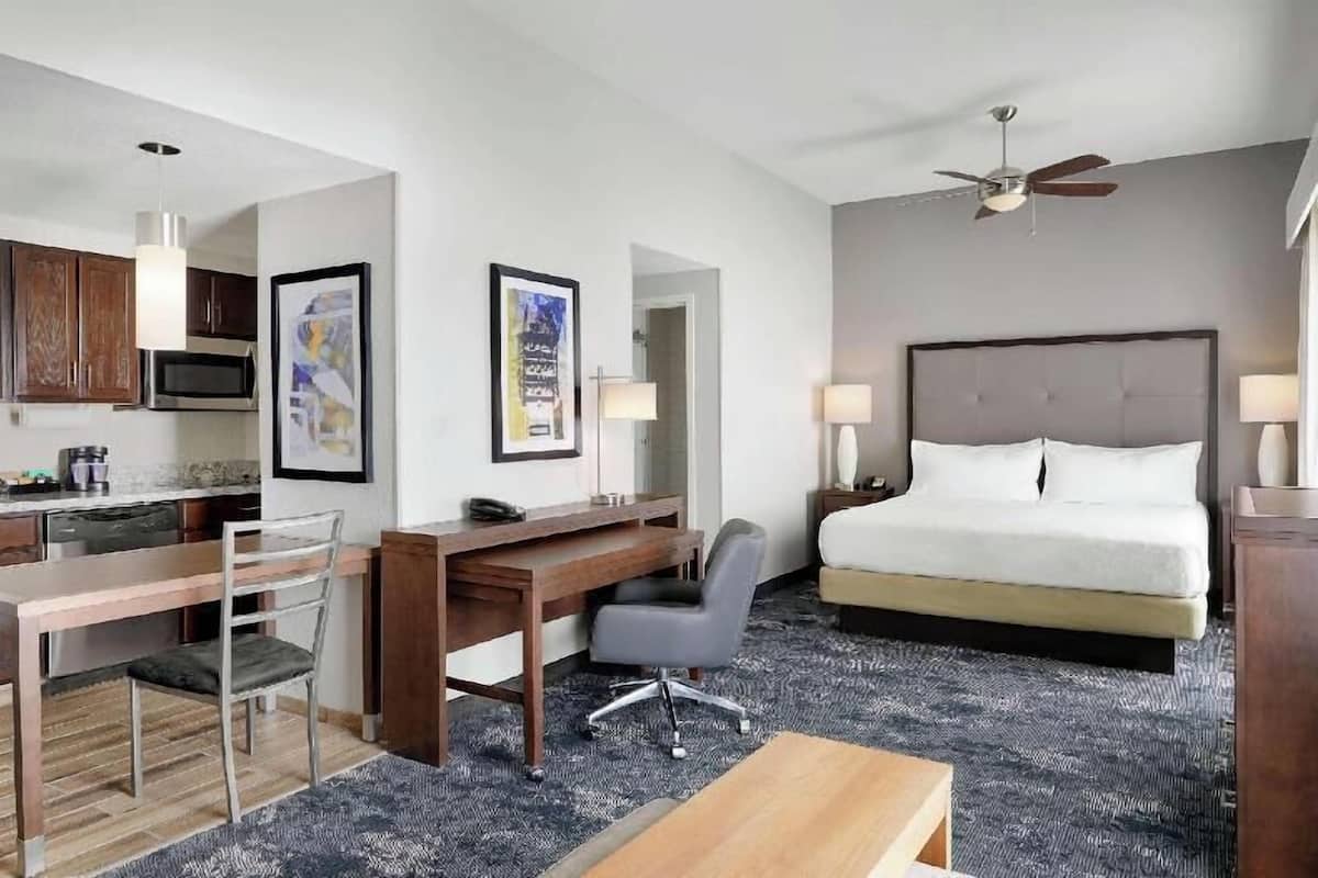 A hotel room with a bed, desk, chair, kitchenette, and artwork.