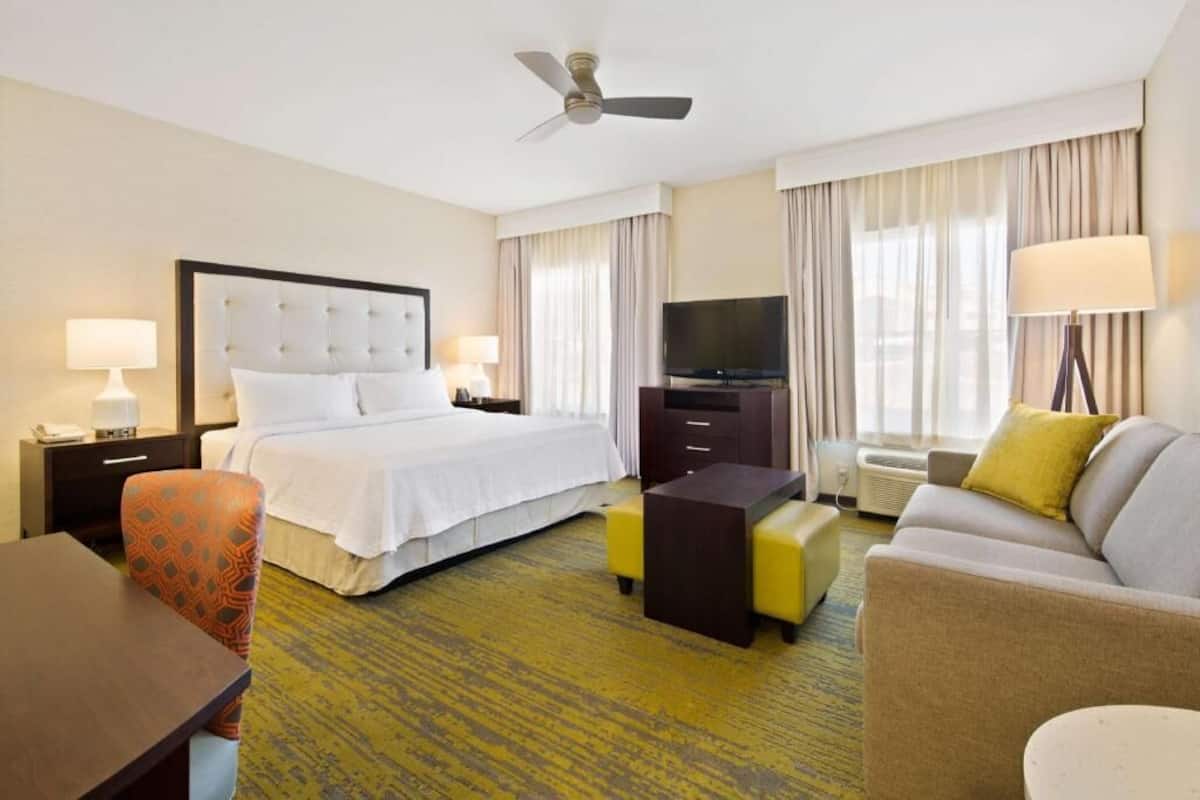 A hotel room with a bed, a television, a desk, a chair, a sofa, and a ceiling fan.