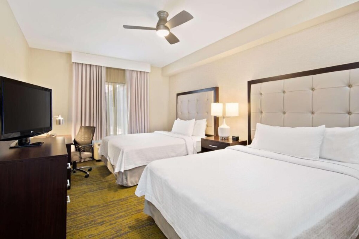 A hotel room with two beds, a television, a desk, a chair, a ceiling fan, and a window with curtains.