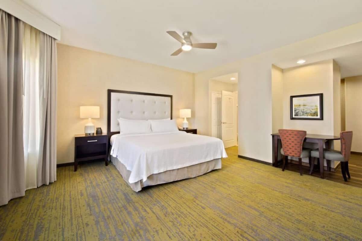 A hotel room with a bed, a desk, two chairs, a ceiling fan, and a door.
