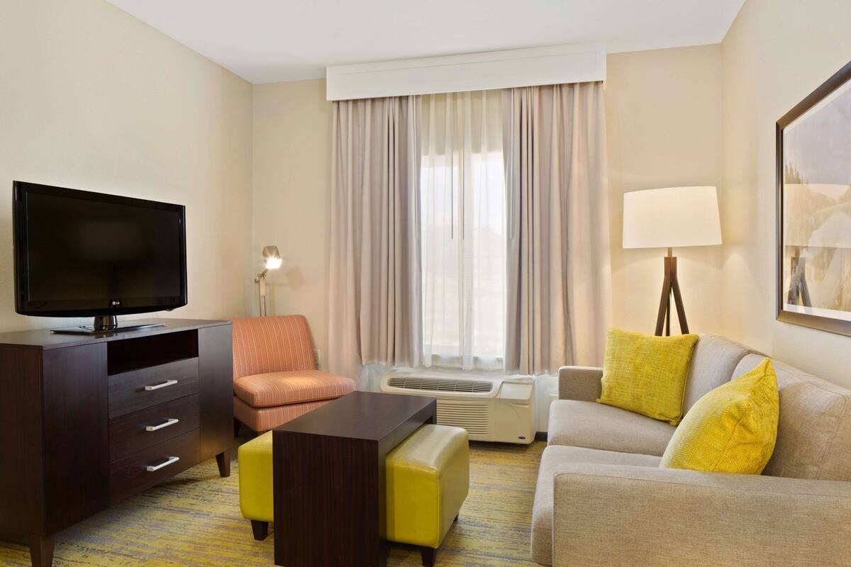 A hotel room with a flat-screen TV, a sofa, a chair, a desk, and a lamp.