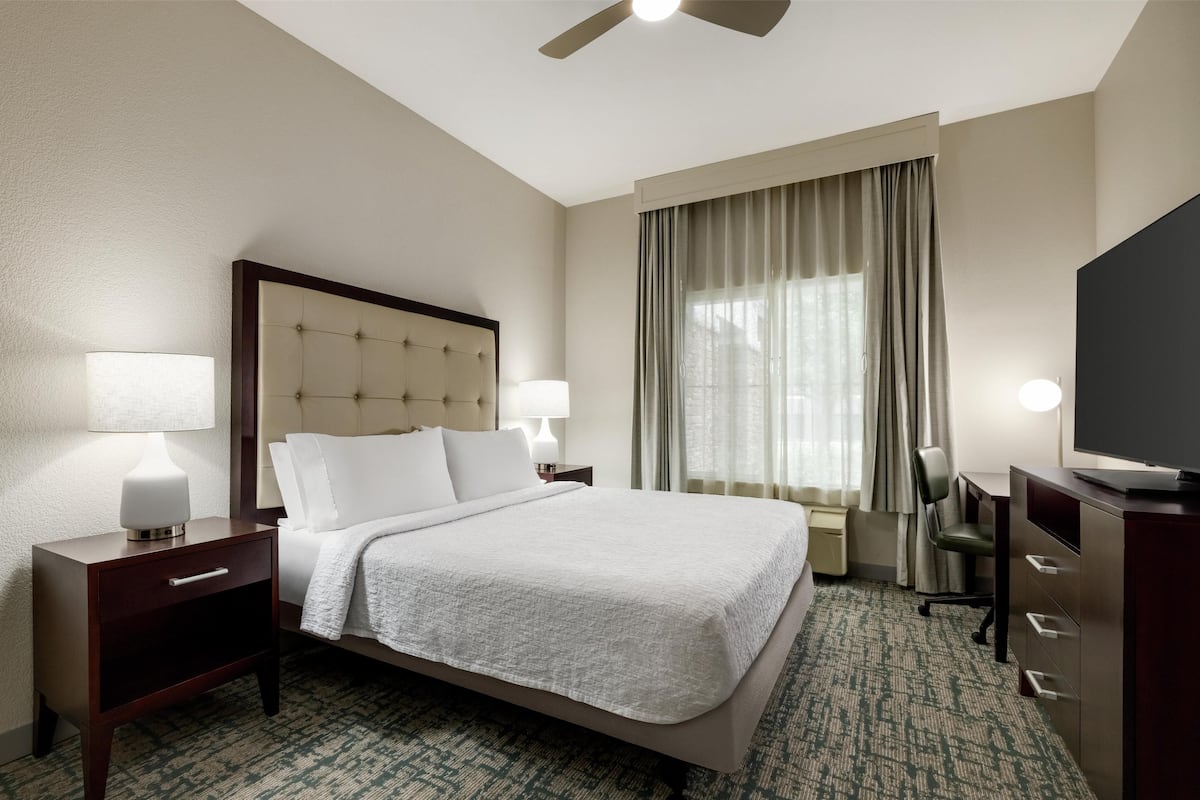 A hotel room with a large bed, two bedside tables with lamps, a desk with a chair, a ceiling fan, and a large window with curtains.