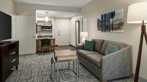 A hotel room with a sofa, a small table, a chair, a TV, a lamp, a painting, and a kitchenette with a microwave. - Homewood Suites by Hilton Denver West Lakewood (Lakewood)