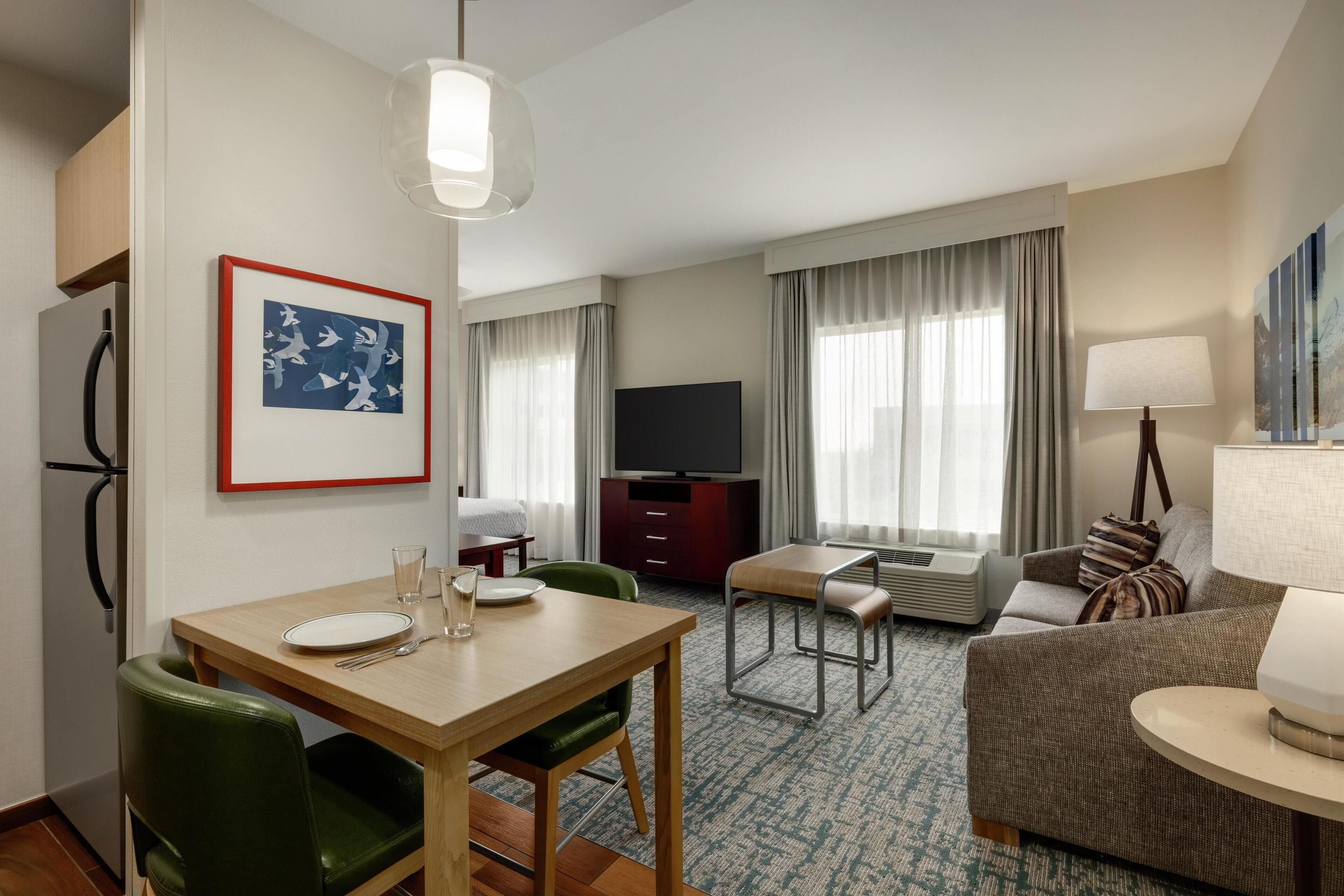 Ett hotellrum med ett matbord, gröna stolar, en soffa, en tv och ett fönster med gardiner.