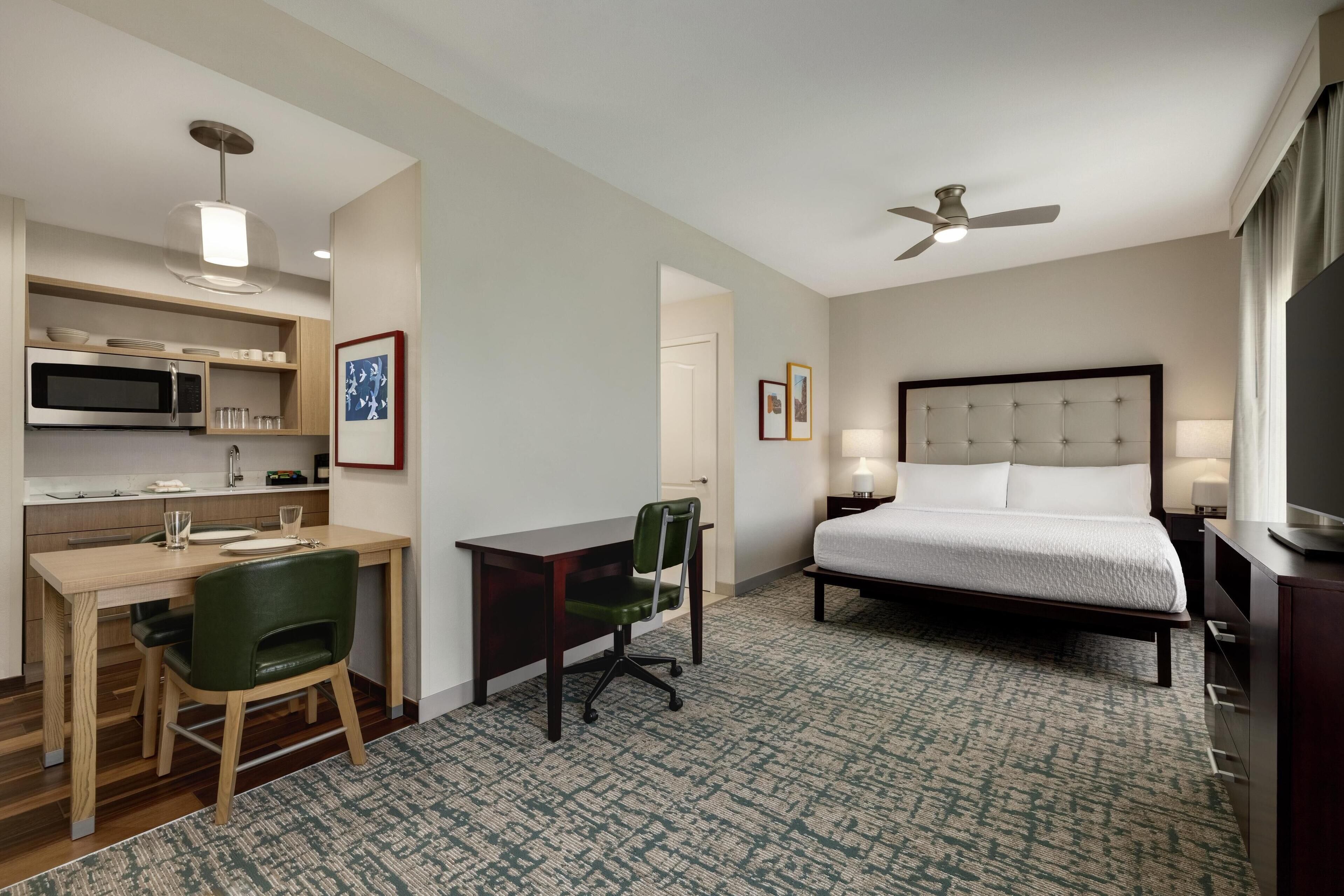 A hotel room with a bed, desk, chair, microwave, and ceiling fan.