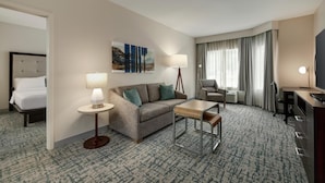 A hotel room with a bed, a sofa, a small table, a lamp, a TV, and a large window with curtains. - Homewood Suites by Hilton Denver West Lakewood (Lakewood)