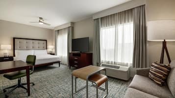 Quarto de hotel com cama, escrivaninha, cadeira, sofá, televisão, luminária e janela com cortinas.