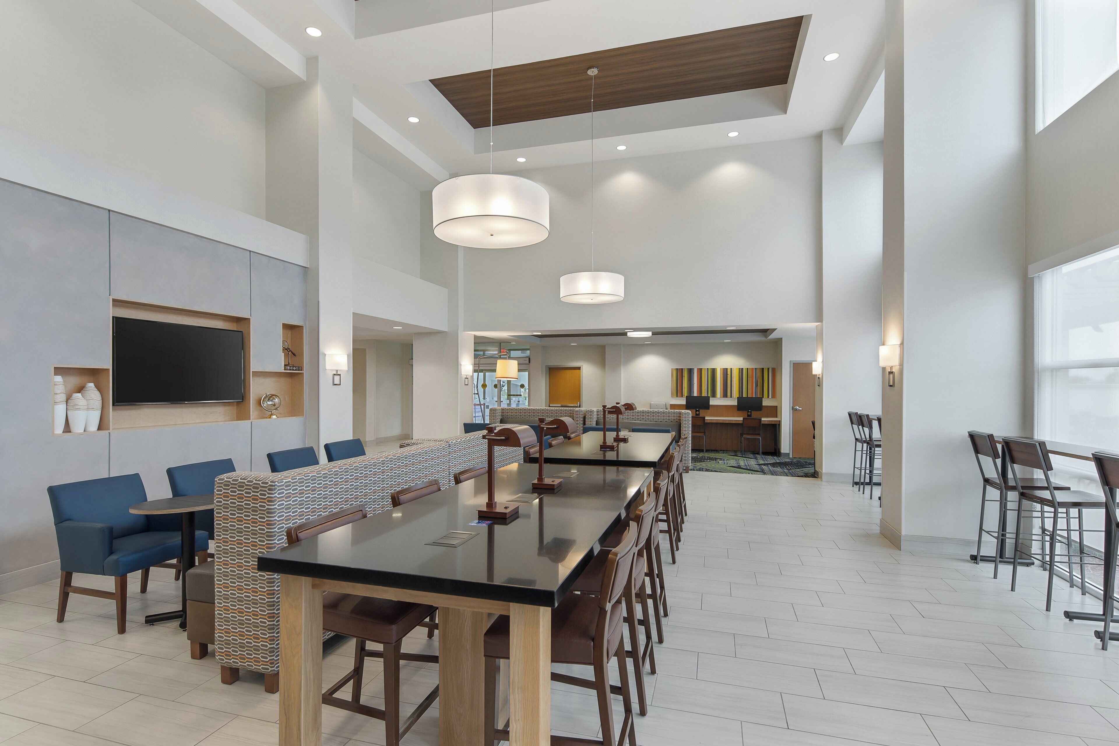 A spacious hotel dining area with a long communal table, multiple seating options, and a modern ceiling design.