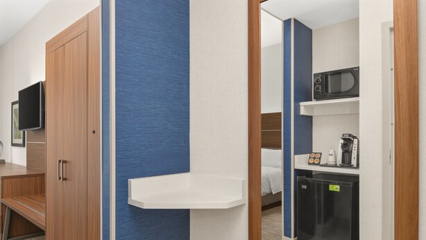 Ein Hotelzimmer mit Holzschrank, Flachbildfernseher, einer kleinen Küchenzeile inklusive Mikrowelle, Kaffeemaschine und Kühlschrank.