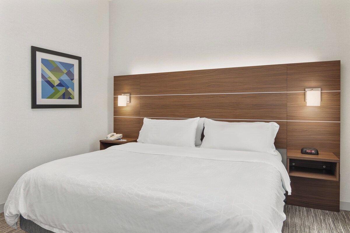 Habitación de hotel con una cama grande, dos mesitas de noche, una lámpara de noche y una obra de arte abstracta enmarcada, además de luces fijadas en la pared.