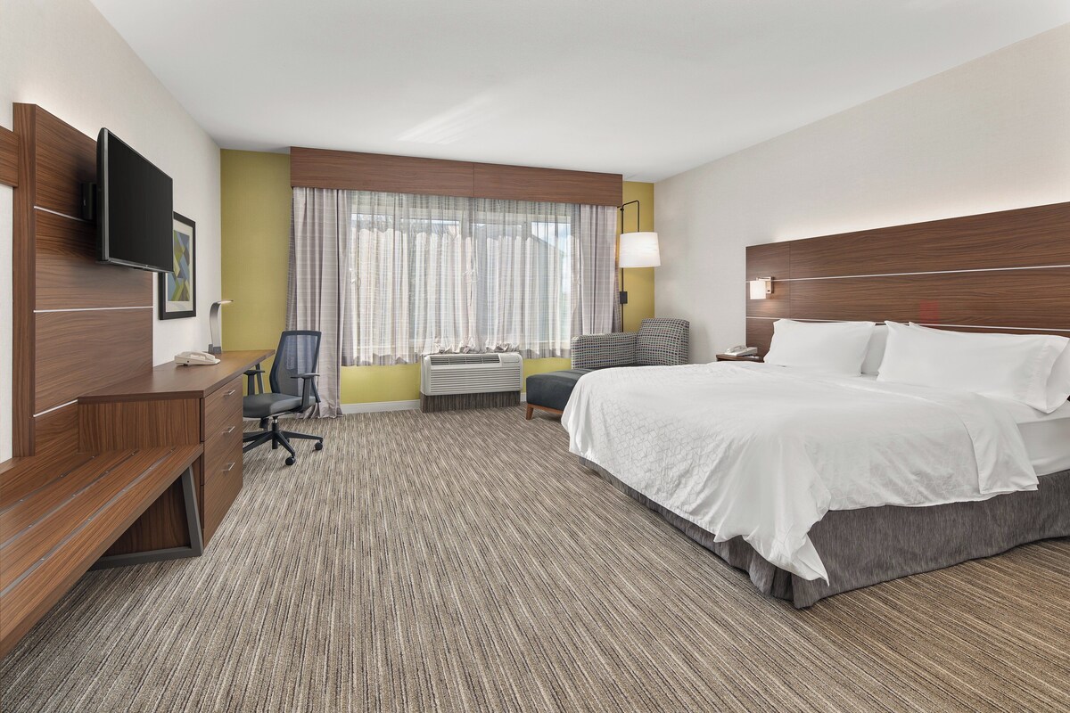 Habitación de hotel con cama, escritorio, silla, televisión y ventanas grandes con cortinas.
