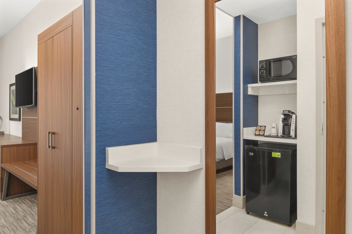 Habitación de hotel con armario de madera, televisor de pantalla plana, estante blanco, cama, refrigerador negro y microondas.