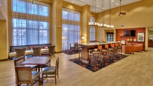 A hotel dining area with a central island, multiple seating options, and large windows with curtains. - Hampton Inn & Suites Fort Myers-Estero/FGCU (Estero)