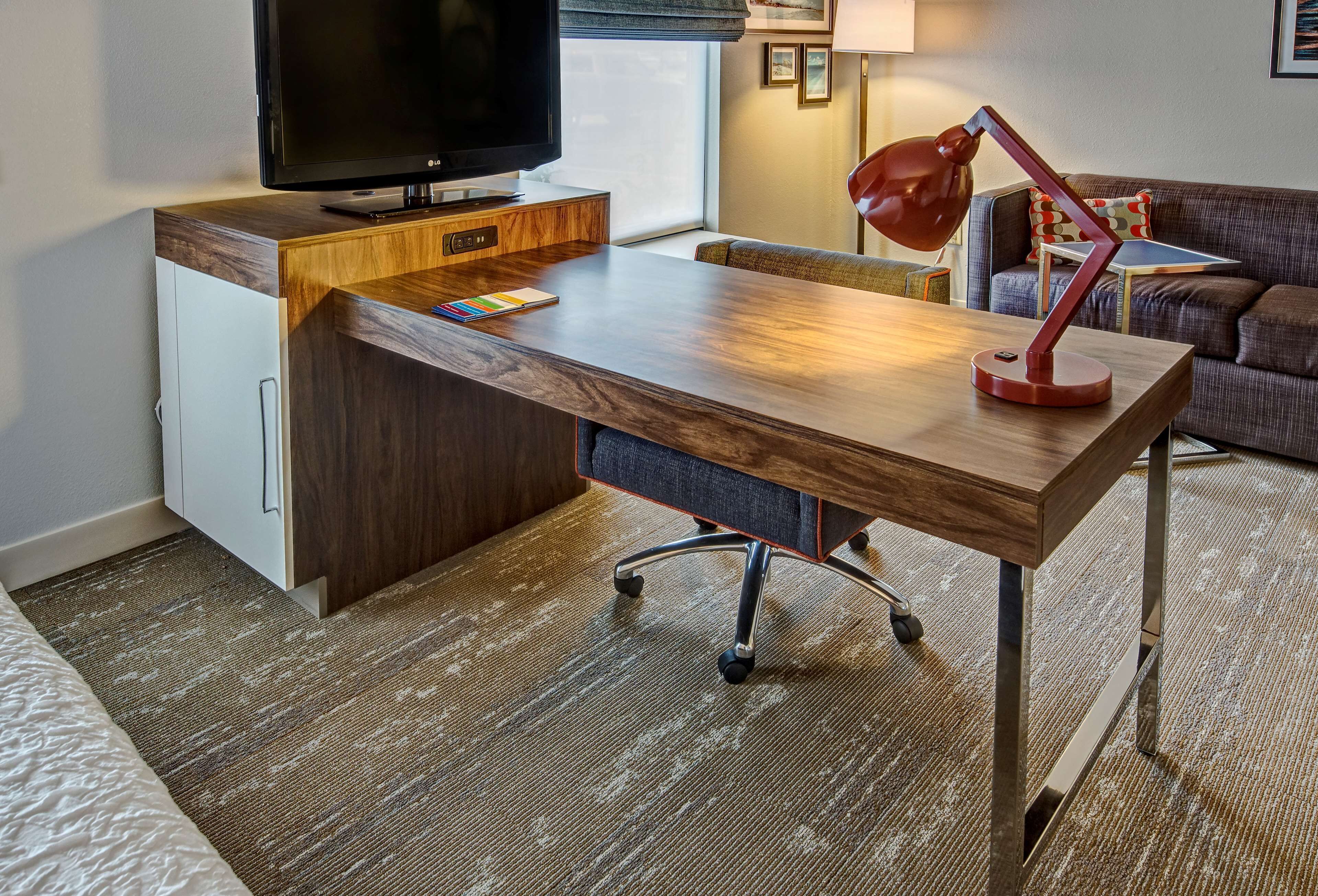 Una habitación de hotel moderna con un escritorio de madera, un televisor, una lámpara de escritorio roja y un sofá.