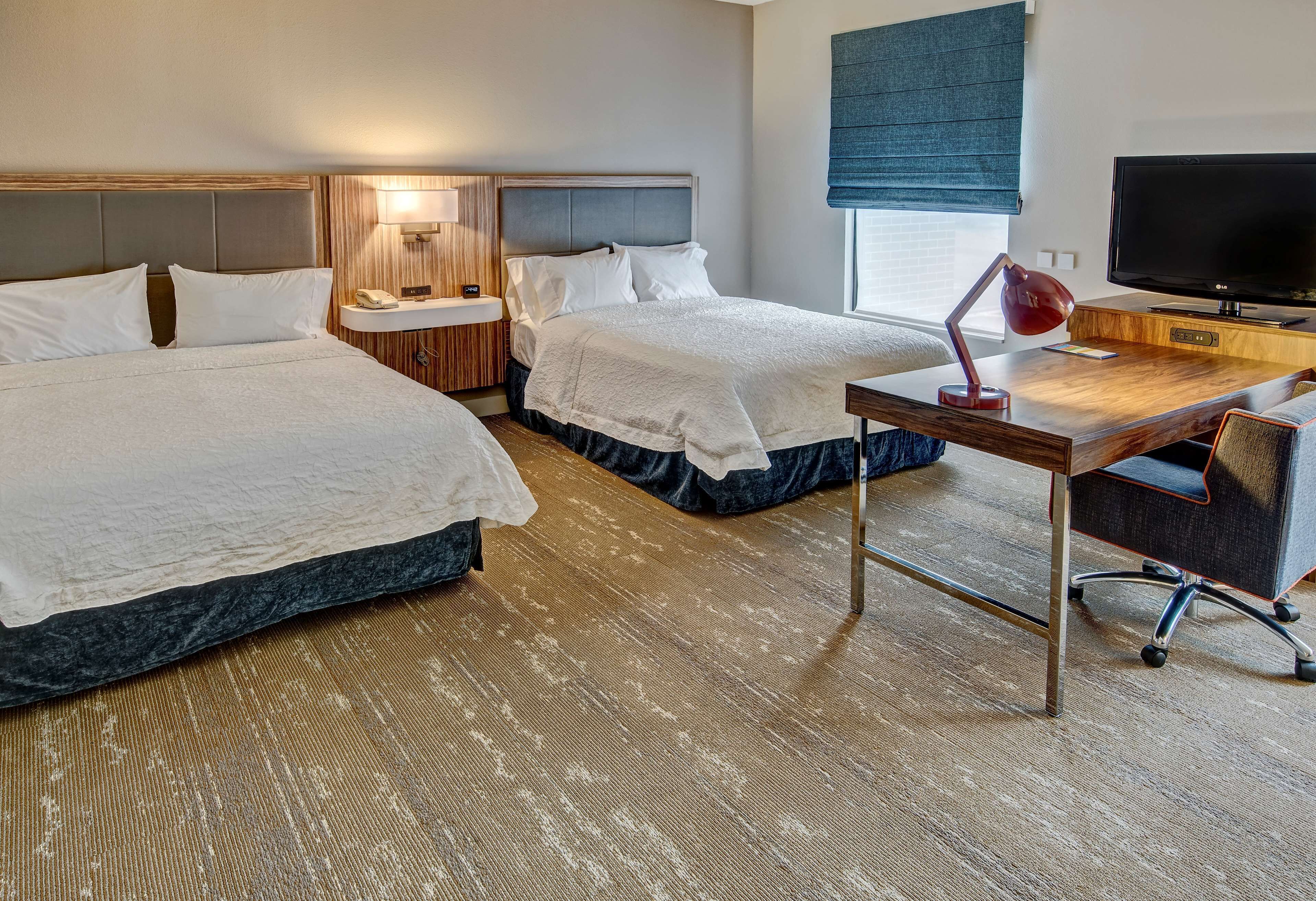 A hotel room with two beds, a desk with a lamp, a TV, and a window with a blue blind.