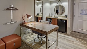 A modern hotel room with a wooden desk, a leather ottoman, a desk lamp, a chair, a large mirror, a sink, and a refrigerator. - Hampton Inn & Suites Fort Myers-Estero/FGCU (Estero)