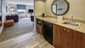 A modern kitchen with a granite countertop, a sink, and a dishwasher. There is a dining area with a table and chairs visible in the background. - Hampton Inn & Suites Fort Myers-Estero/FGCU (Estero)