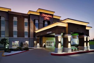Entrada de um hotel Hampton Inn com um letreiro 'Welcome', um acesso coberto e área ajardinada com arbustos e uma palmeira.