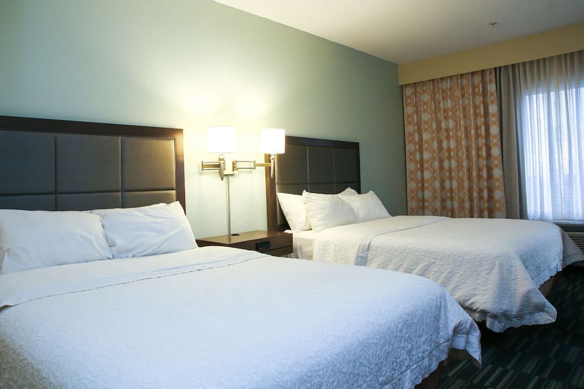 A hotel room with two beds, a nightstand with lamps, a window with curtains, and a headboard.
