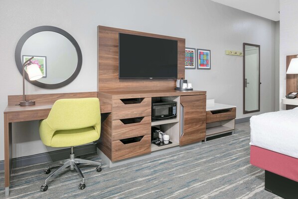 A hotel room with a wooden desk, a yellow office chair, a flat-screen TV, a microwave, a bed with a red and white duvet, a mirror, and a door with a nameplate.