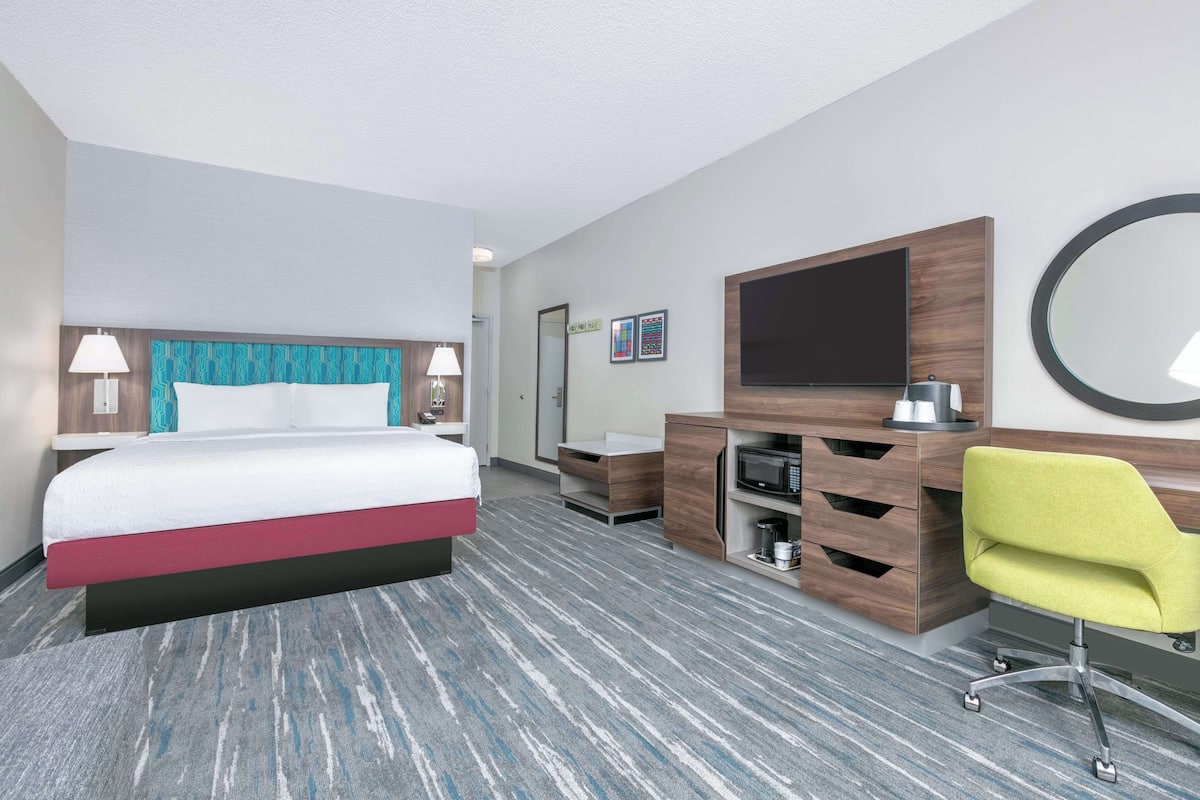 A hotel room with a bed, a desk, a chair, a TV, a mirror, and a microwave.