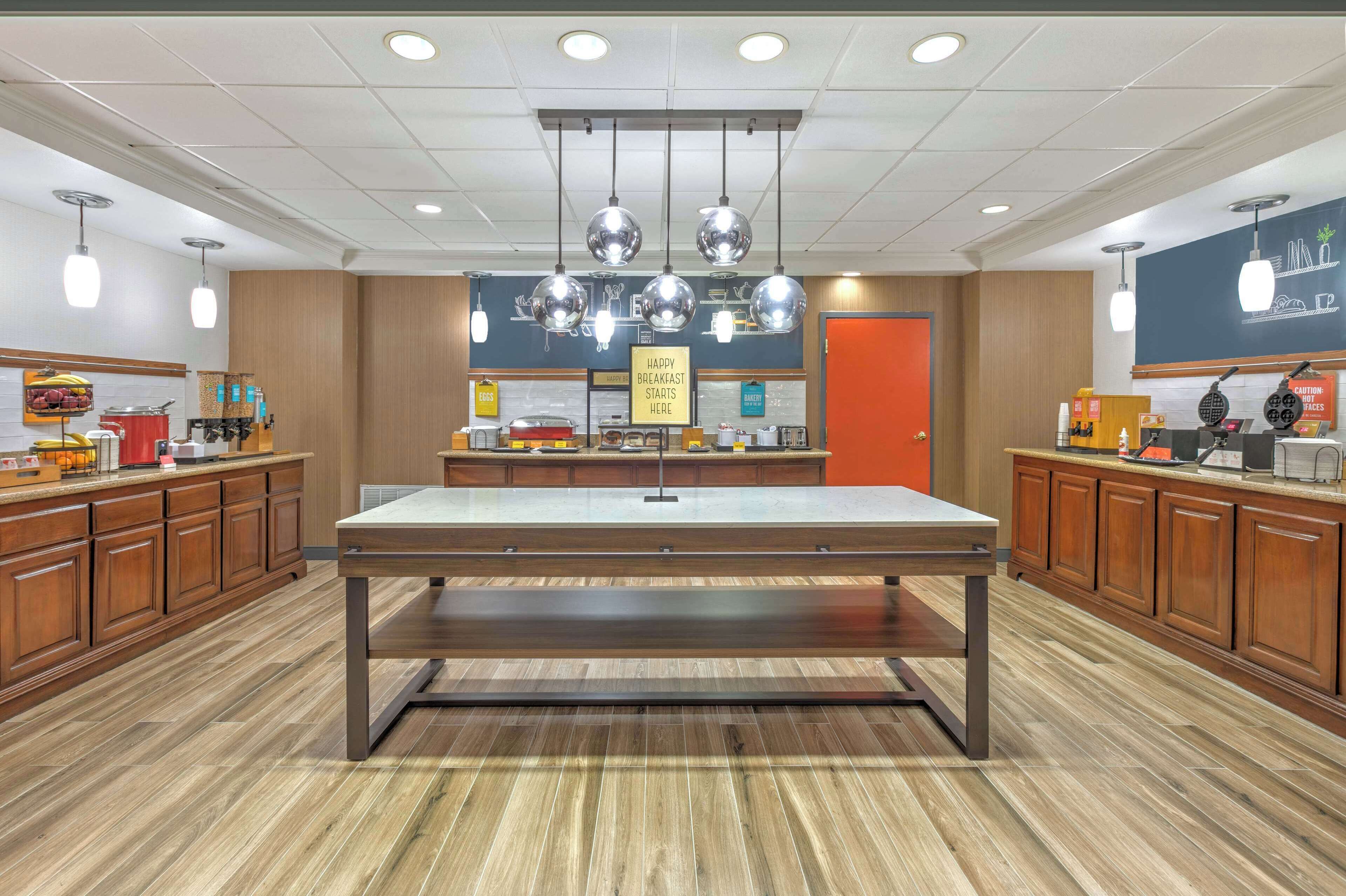 A modern breakfast area with a central table, wooden cabinets, and hanging pendant lights.