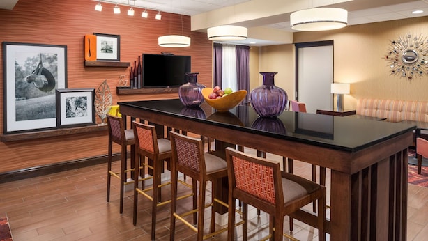 A hotel room with a wooden floor, a large central table with bar stools, a flat-screen TV, and a decorative wall with framed pictures and a mirror. - Hampton Inn Belle Vernon (Belle Vernon)