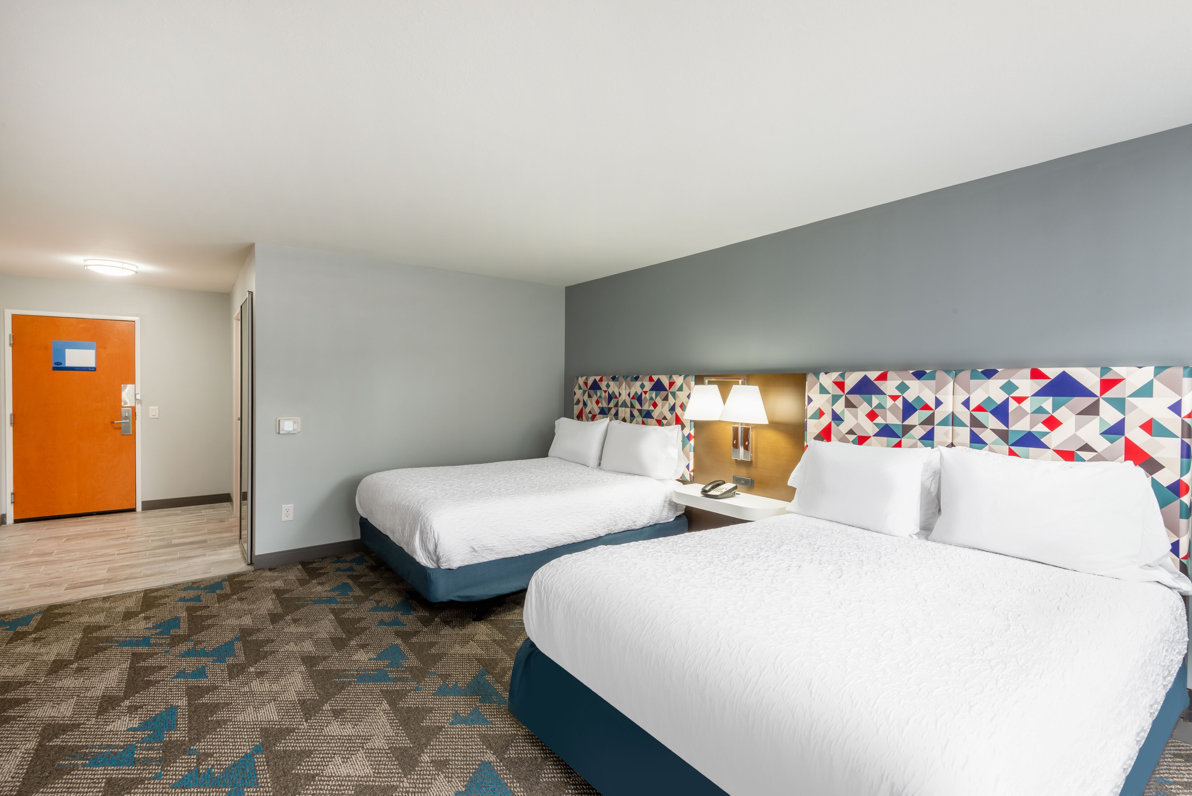 A hotel room with two beds, a patterned carpet, a geometric-patterned headboard, and a door with a sign.