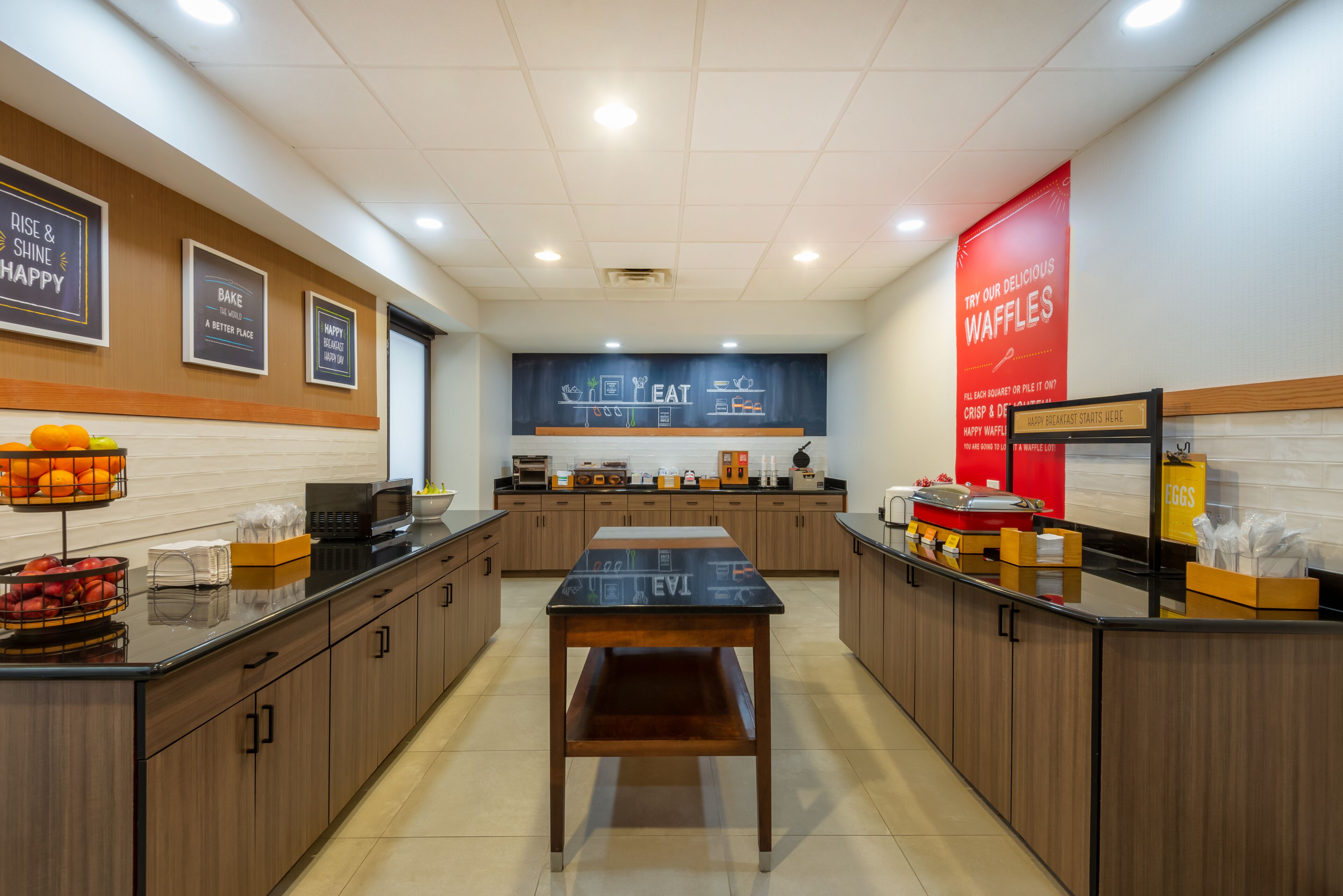 Een moderne keuken met houten kastjes, een zwart aanrecht en een centrale tafel. Op de muren staan borden met aanbiedingen voor wafels en ontbijtartikelen.