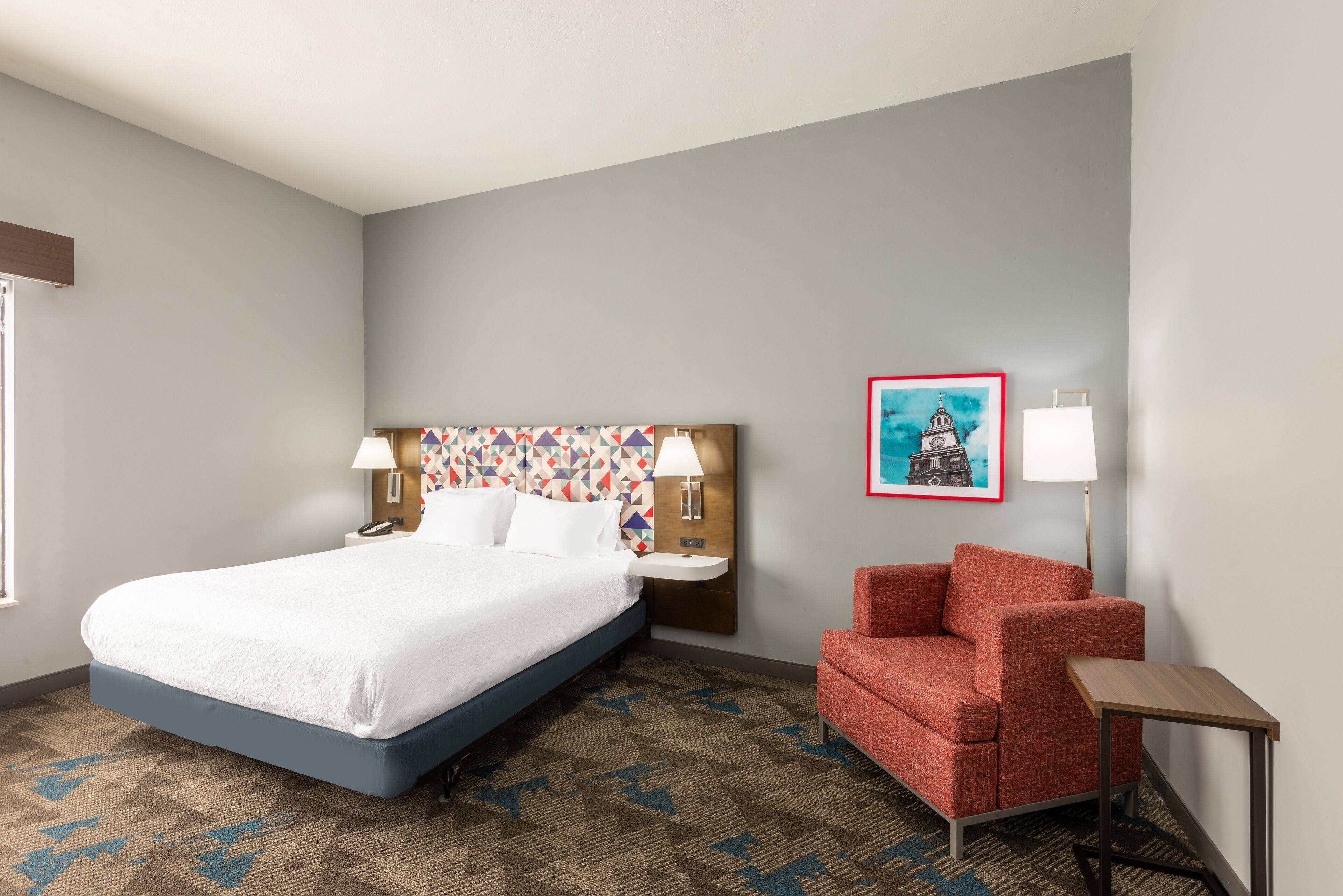 A hotel room with a bed, a red chair, a nightstand, a lamp, and a painting on the wall.